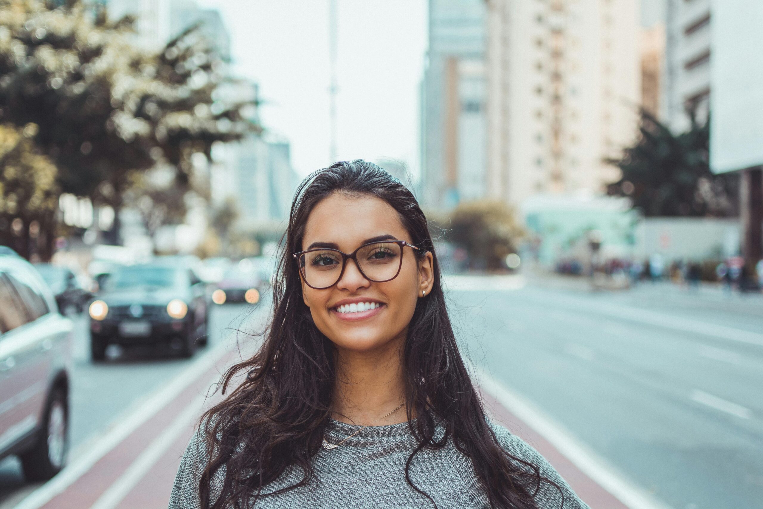 Home A young woman with glasses smiling on a city street, embracing urban lifestyle.